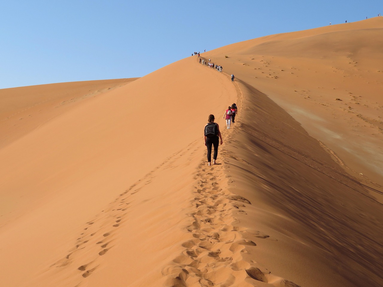sur la crête d'une dune de sable nous voyons des personnes qui marchent en file indienne vers une même direction. Les traces de leurs pas sont marquées dans le sable